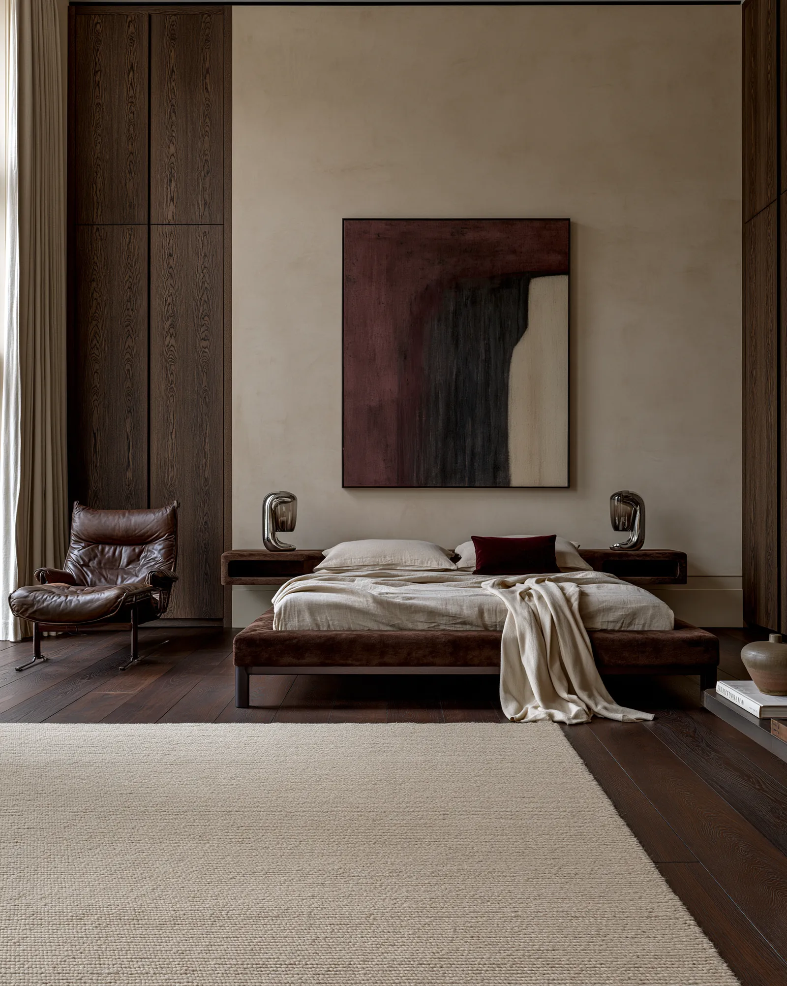 Bedroom with a brown velvet bed, floating timber bedside shelves and chrome table lamps beneath a dark red abstract canvas, with a vintage leather lounge chair and ottoman