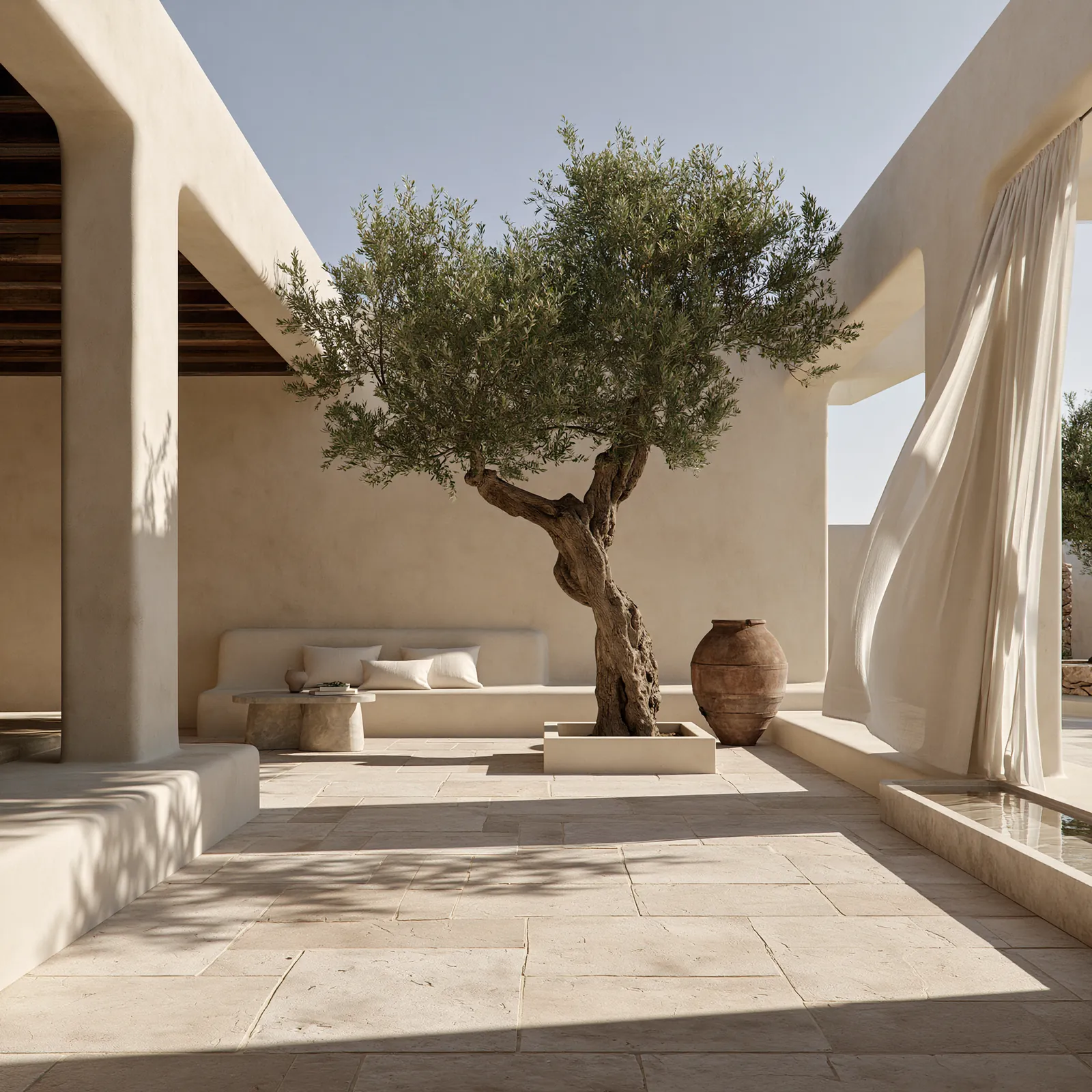 Enclosed courtyard with a built-in plaster bench, a stone table and an olive tree in a low planter, beside a narrow water channel and a linen curtain