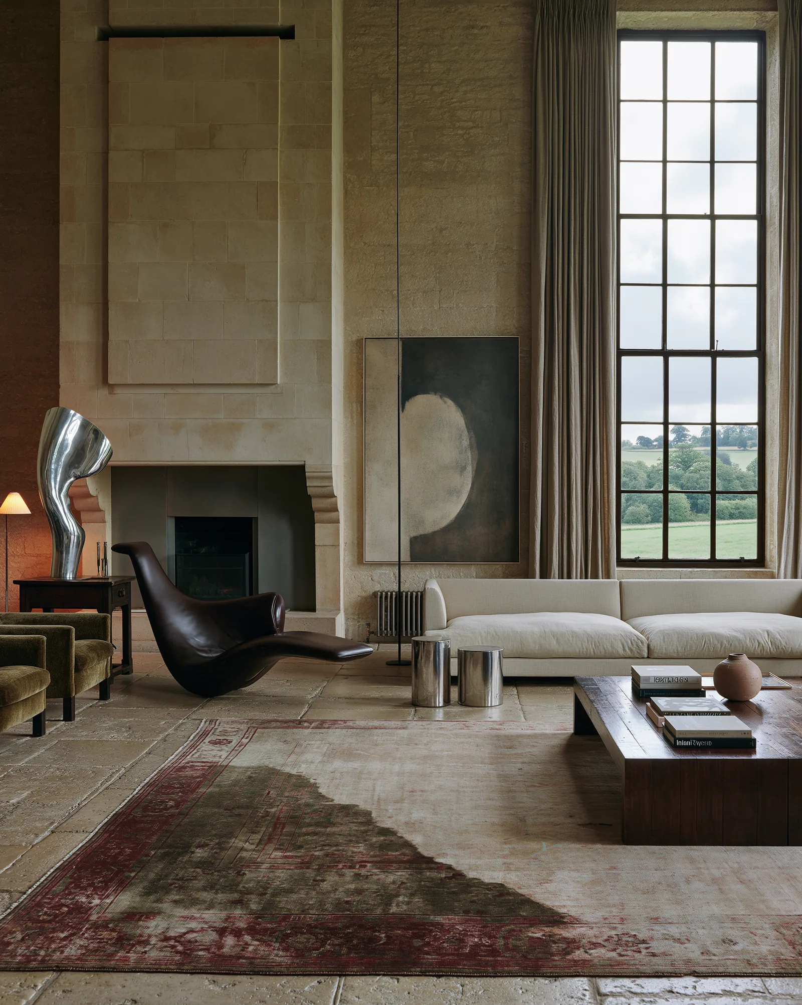Principal drawing room with a monumental limestone fireplace, a tall mullioned window onto open countryside, a long ivory sofa and a dark leather chaise