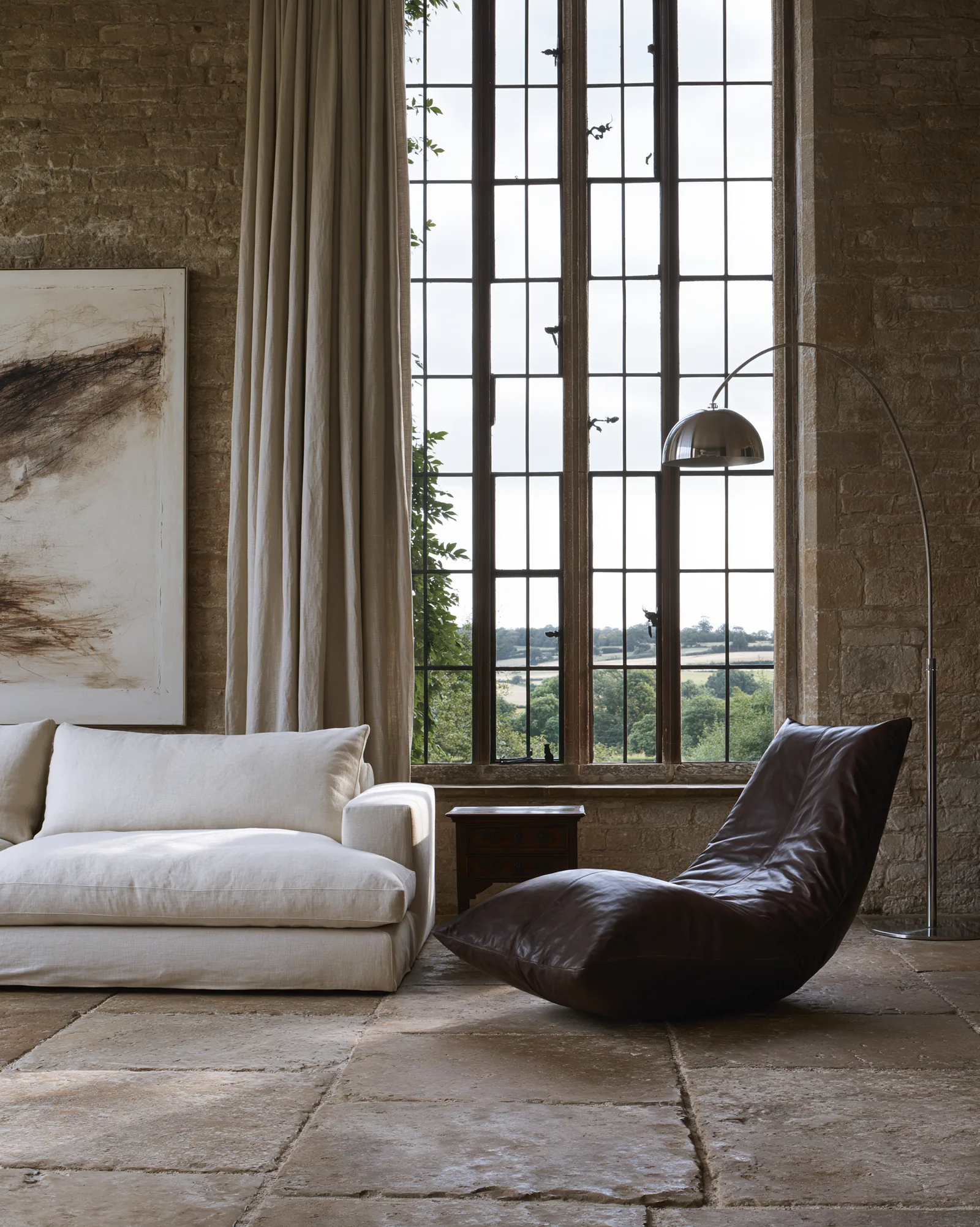 Sitting room against exposed stone, with a linen sofa, a dark leather chaise and an arc lamp beneath a full-height window