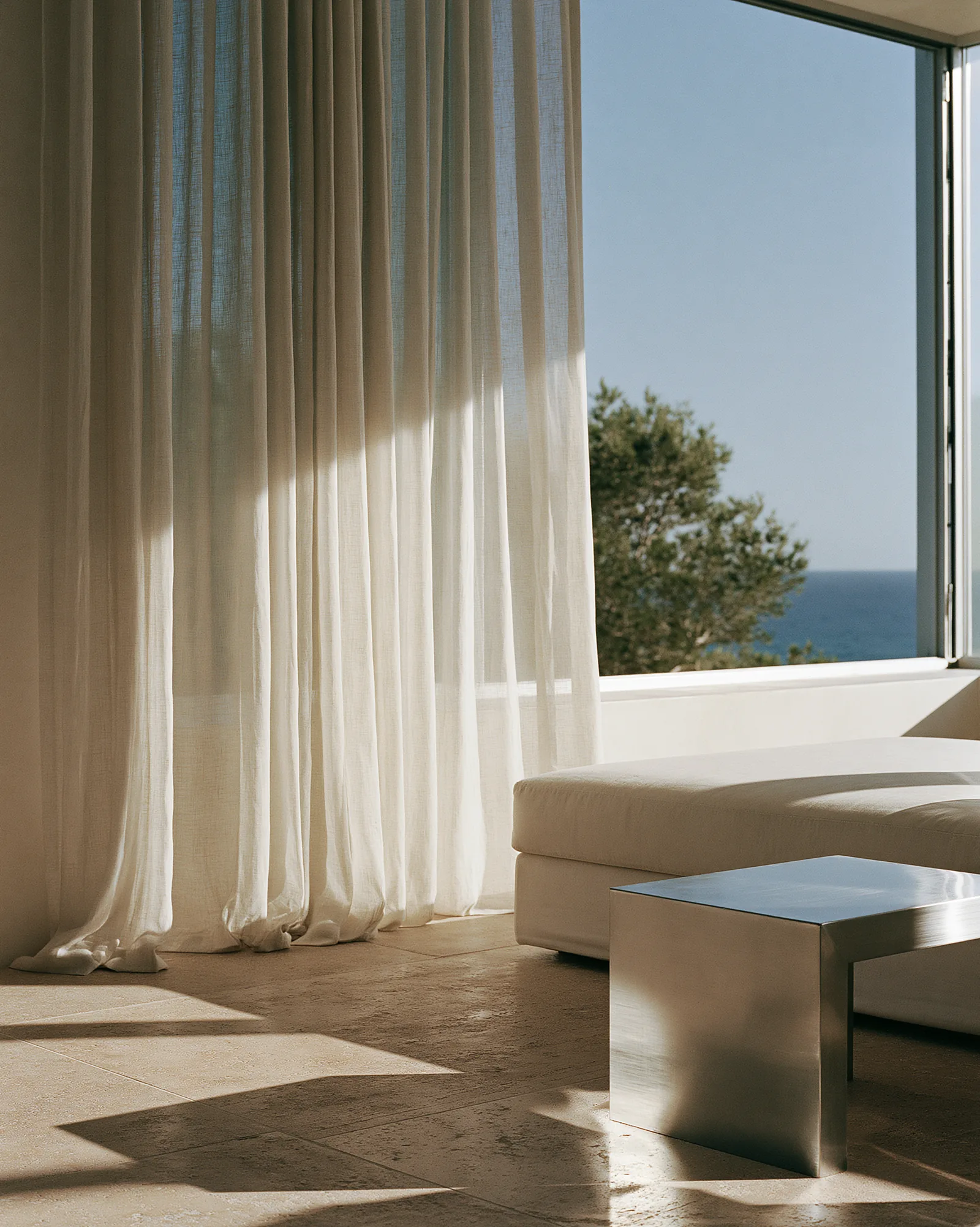 Son Vida House — living room opening to the Mallorcan coastline through full-height linen curtains, ivory seating and a polished steel table on pale stone