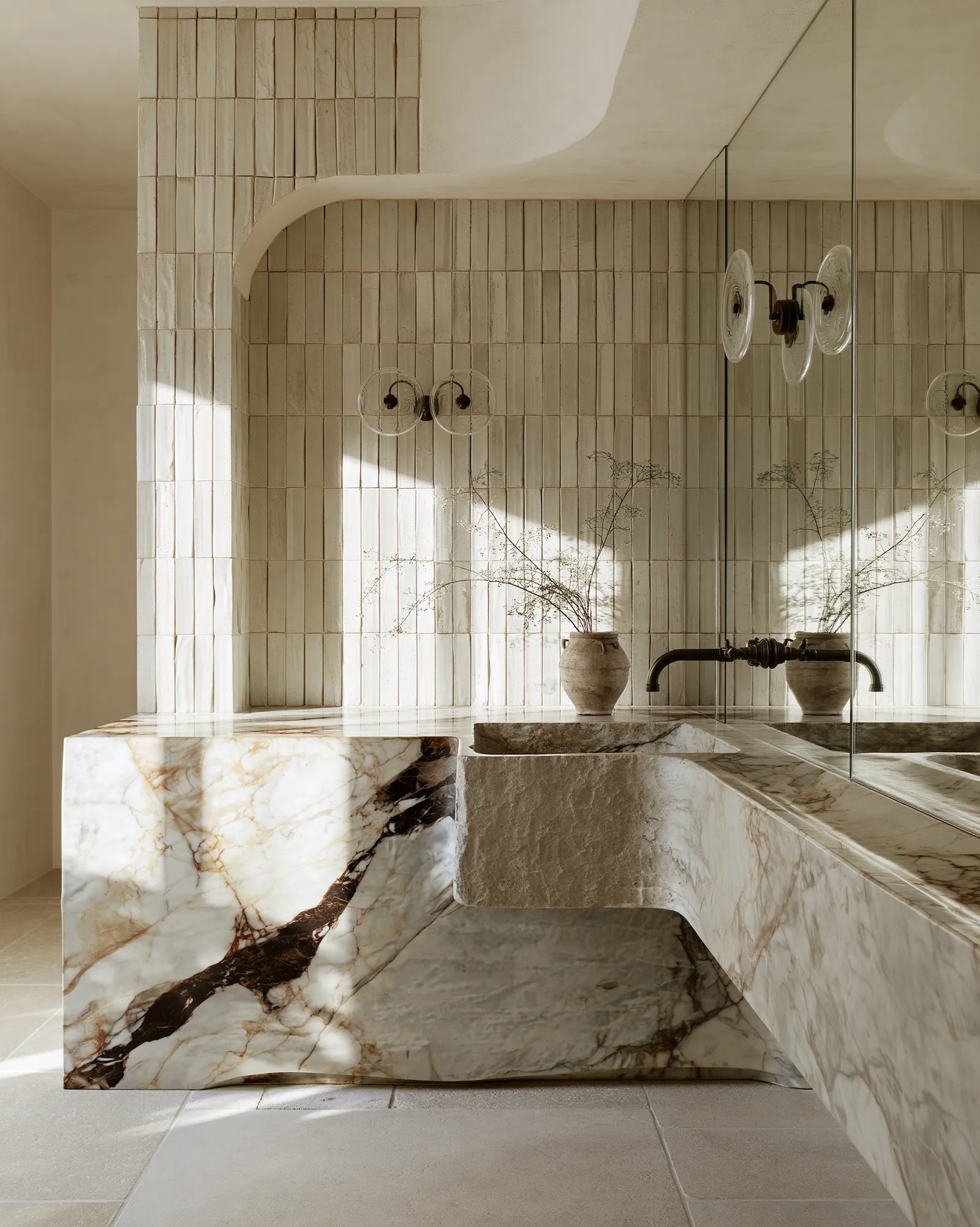 Baño con lavabo tallado en piedra sobre encimera de mármol veteado contra azulejo vertical esmaltado a mano, con grifería de metal envejecido y una tinaja de barro