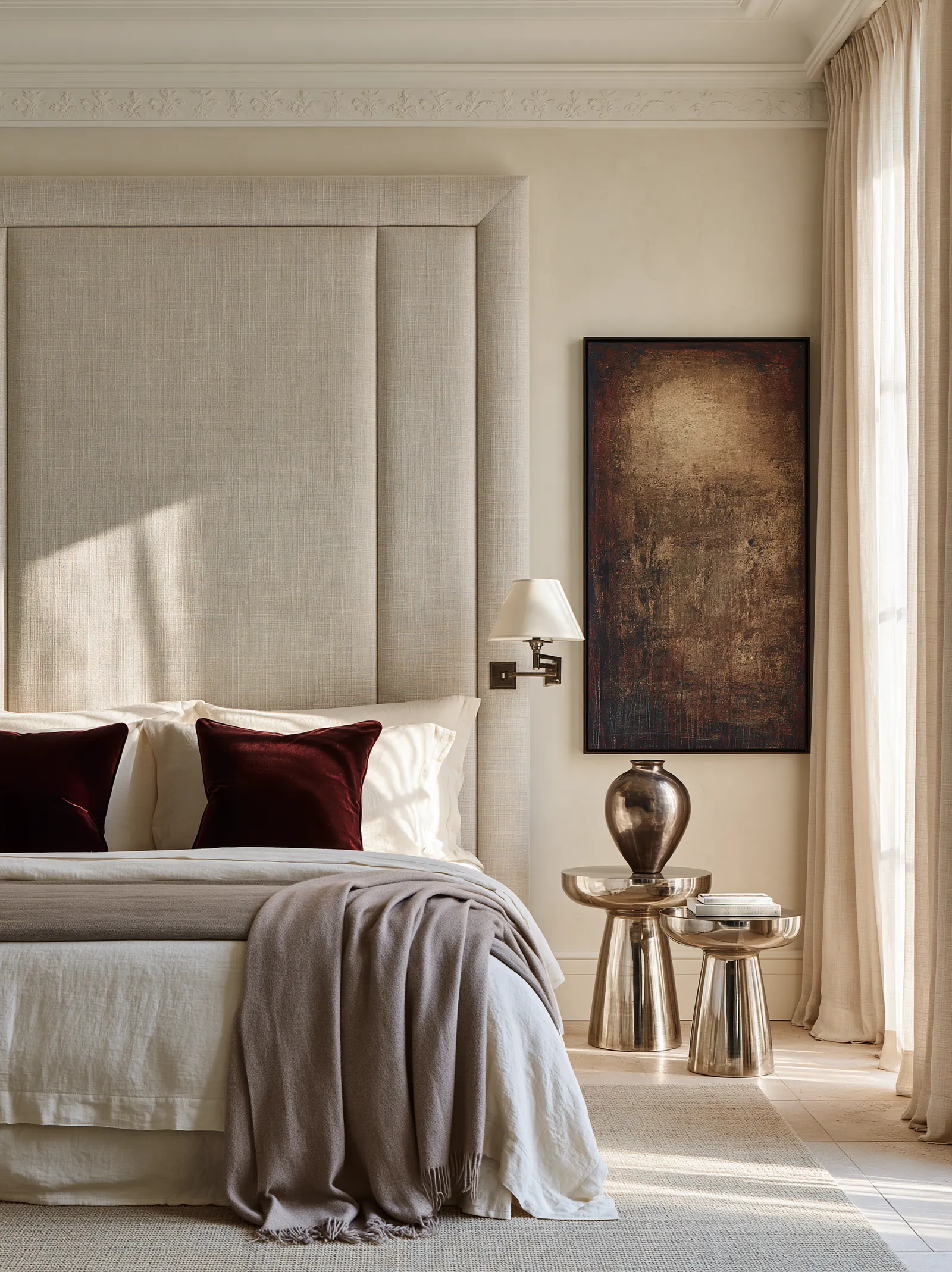 Bedroom with a full-height linen headboard panel, oxblood velvet cushions and polished metal bedside tables beneath a dark bronze canvas