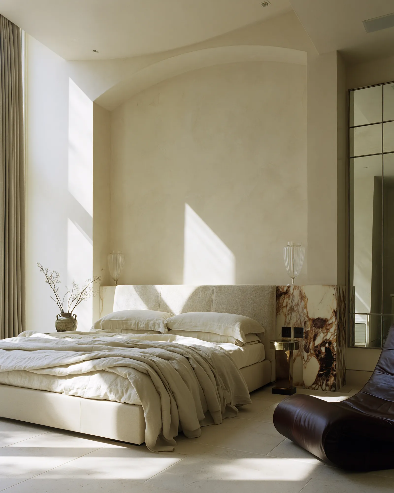 Second bedroom scheme set into a curved plaster alcove, with a bouclé headboard, a veined marble return and a dark leather lounger on limestone