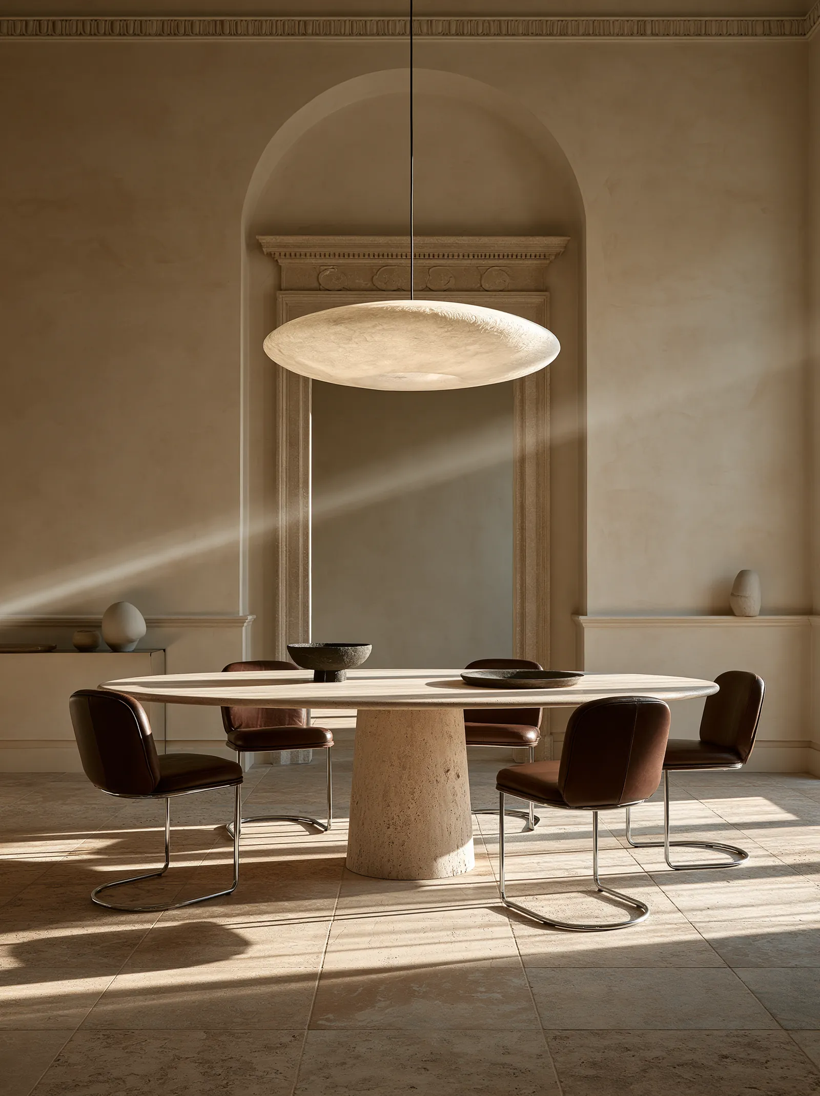 Dining room with an oval travertine table on a conical base and dark leather cantilever chairs, beneath an alabaster pendant and an arched stone niche