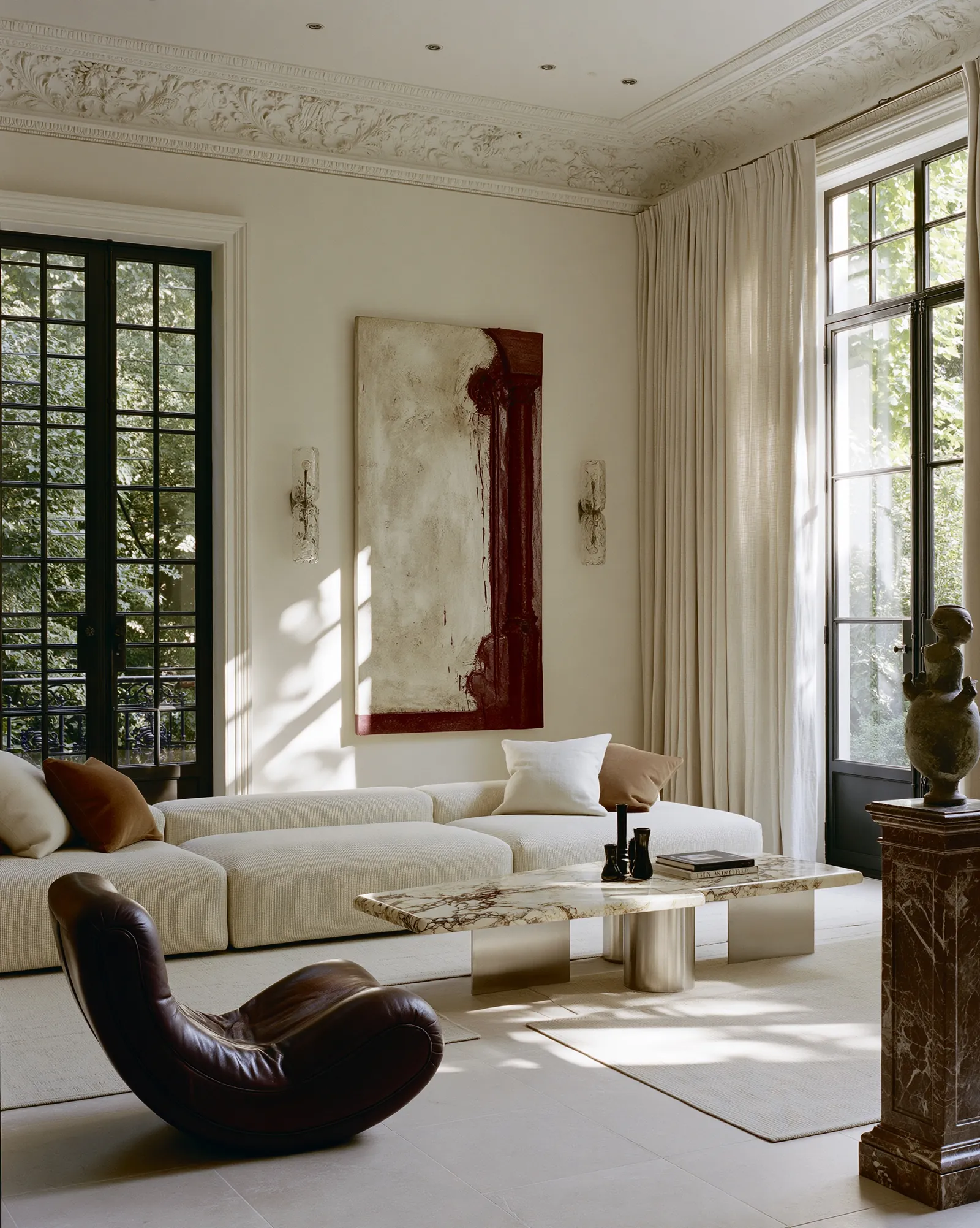 Living room with a deep burgundy canvas above a low linen sofa, a dark leather lounge chair and a veined marble table on limestone, beneath ornate plaster cornicing
